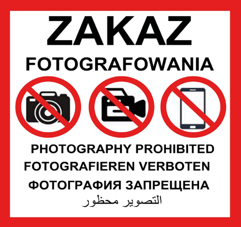 Wzór tablicy informacyjnej. U góry duży czarny napis „Zakaz fotografowania”. Poniżej trzy czarne ikony: aparat fotograficzny, kamera wideo i smartfon, każda przekreślona czerwonym okręgiem z ukośną linią. Pod ikonami napisy w różnych językach: po angielsku „Photography prohibited”, po niemiecku „Fotografieren verboten”, po rosyjsku „Фотография запрещена” oraz po arabsku „التصوير محظور”.
