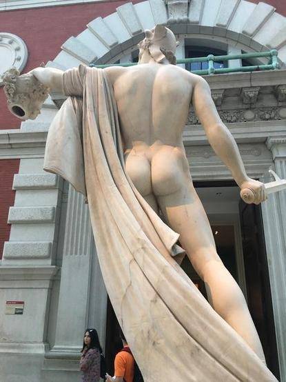 Neoclassical marble sculpture of Perseus holding the severed head of Medusa in his left and his sickle sword in his right. He is shown from reverse, his buttocks and back muscles well-defined in the artwork. He is nude, the cap of invisibility and a long himation draped over his left arm the only clothing.