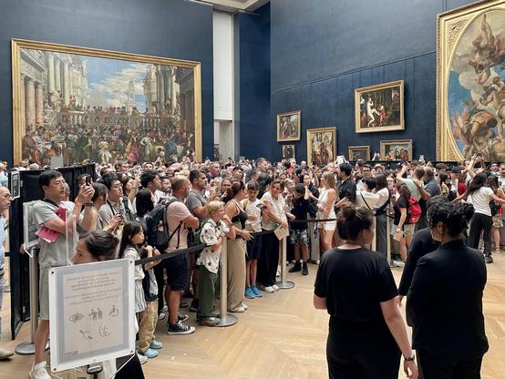 A large crowd of visitors is gathered in an art museum, with people taking photos. Several famous paintings are displayed on the walls, and there are visible queue barriers and signs. The atmosphere is vibrant and busy, characteristic of a popular tourist attraction.