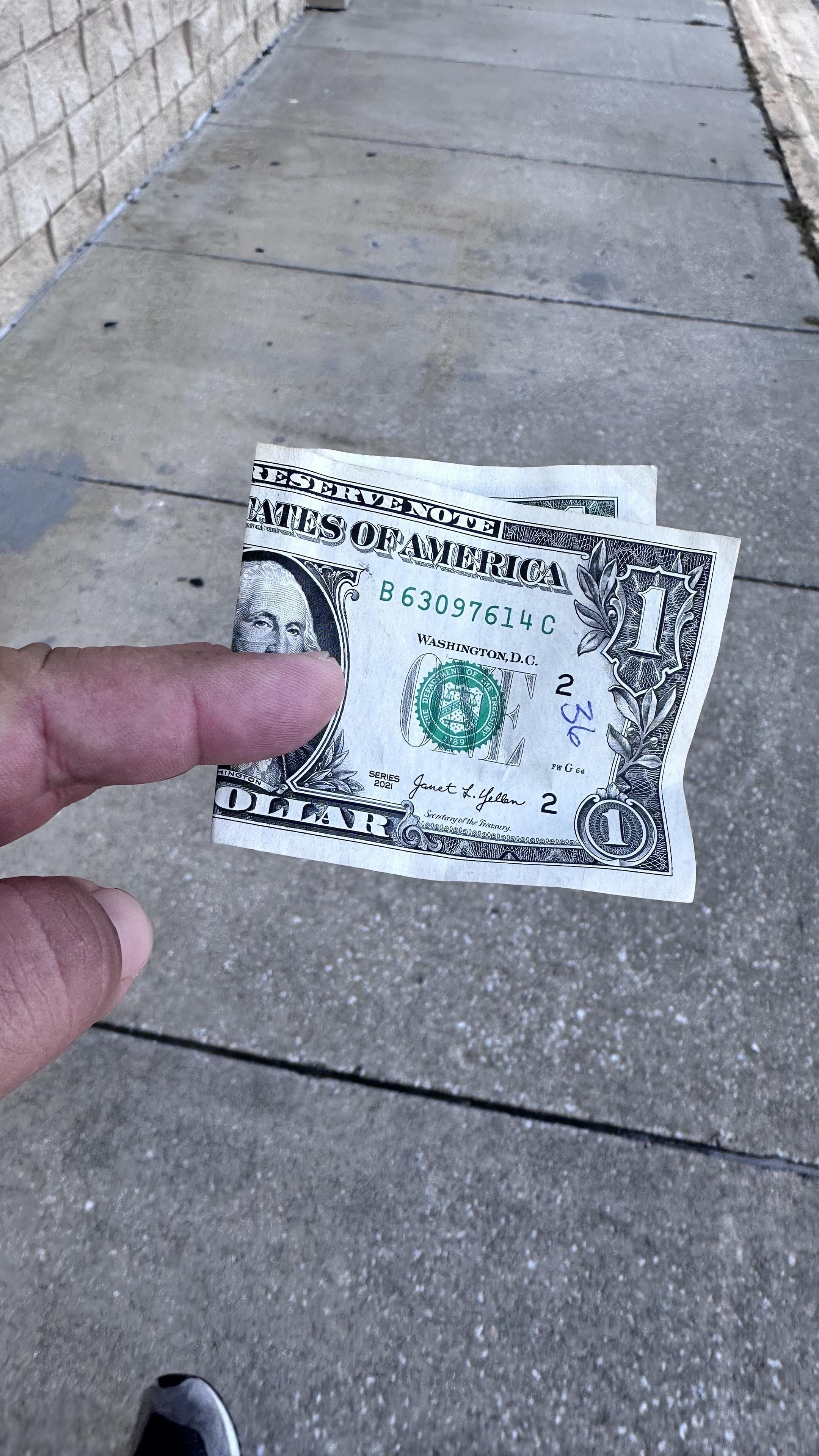 A close-up of me hand holding a one-dollar bill, with a sidewalk in the background. The bill is partially crumpled and shows details like the series date and serial number.
