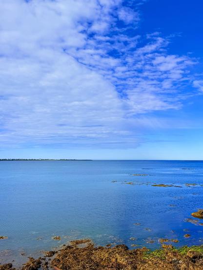 A view out to the water. The left side of the image has clouds and the right mostly blue skies. The ocean reflects this with a light blue color under the clouds and a deeper blue under the sky. 