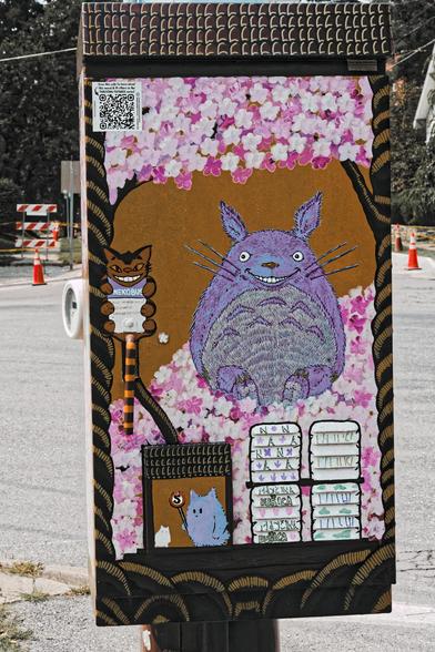 Decorated electric box seen from the long side,  at the very top there is a brown stripe with 4 rows of white L marks,  then a field of Sakura petals with a small QR code in the corner then a section of brown background,  though both sounds are surrounded by tree trunks wrapped in spirals,  there's a sign for a nekobus stop and a big totoro character standing in a big pile of fallen sakura petals and below some panels of symbolic characters and a little image of a similar but smaller electric b…