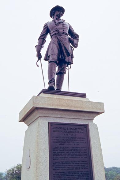 Looking up on a tall pillar to a statue of a man wearing a hat with a wide brim and a double breasted jacket and I think two swords against a light grey sky.
