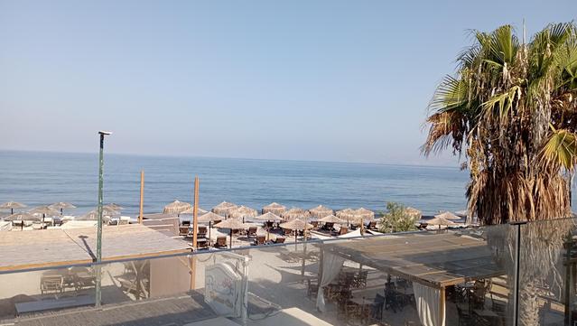 eine idyllische Küstenlandschaft mit einem klaren blauen Himmel und ruhigem Meer. Im Vordergrund Balkon mit gläsernen Trennwänden, die einen Blick auf die Strandbereich ermöglichen. Es gibt mehrere Sonnenschirme aus Stroh, die über Liegestühlen aufgestellt sind, die zum Entspannen einladen. Ein große Palmenbaum steht rechts im Bild und verleiht dem Ort eine insgesamt mediterrane Atmosphäre. Der Strandbereich erstreckt sich bis zum Horizont, wo das Meer in den Himmel übergeht. Die Szene wirkt ru…