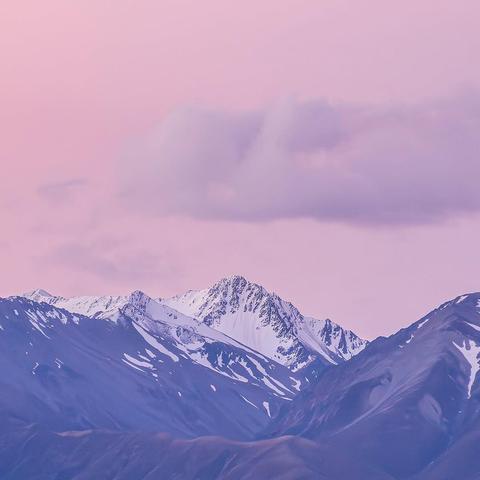 Snow-capped mountain peak in Fiordland National Park, New Zealand, seen under a pink and purple sky. The rugged terrain is surrounded by alpine ridges and dramatic light, evoking the remote beauty of the South Island.