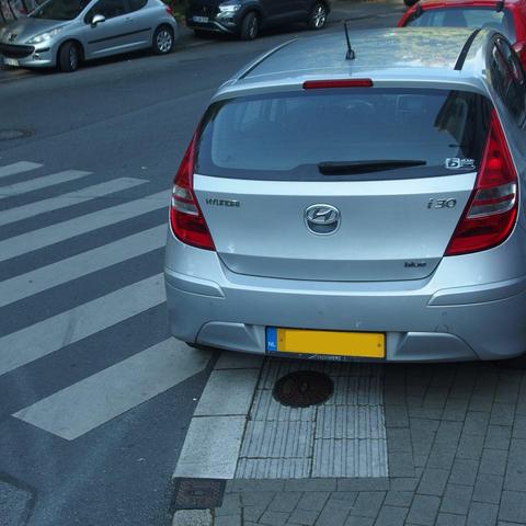 PKW mit niederländischem Kennzeichen falsch-parkt auf dem Gehweg direkt auf einem Fußgängerüberweg. Das Fahrzeug verdeckt auch den Großteil eines Bodenleitsystems.