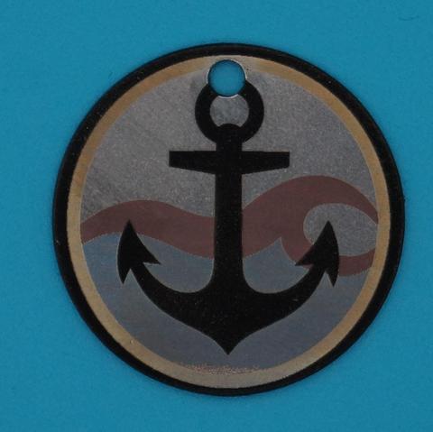 Runder Edelstahlanhänger mit graviertem schwarzen Anker vor wellenförmigem Hintergrund, goldfarbener Rand, auf blauem Untergrund