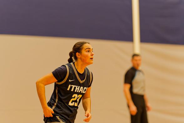 A woman with her hair tired back wears a blue jersey that reads ITHACA 23 and in the background a blurry ref look ons and the wall behind is white below and blue on top