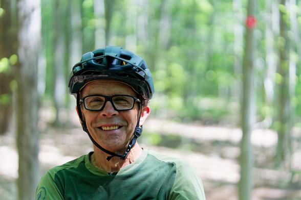 Older gentleman with a blue bike helmet,  thick-rimmed glasses, a broad smile and a two-toned green shirt with blurred out tree stems and green foliage in the background not to mention a red circular trail market