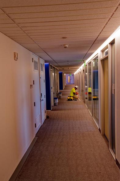 Hallway with a brown carpet on the floor,  a dropped ceiling with small panels,  a mostly white wall on the left interrupted by a conference room about halfway to the end,  on the right find a number of offices with glass fronts, at the end of the hallway is a purplish-blue wall with I think a yellow door and green glowing exist sign.  A man with a white hardhat, yellow shirt and yellow bag is sitting across from the conference room and working on the front of an office on the other side.