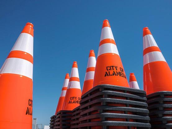 Stacks of orange traffic cones with "City of Alameda" stenciled on them, against an empty blue sky.
