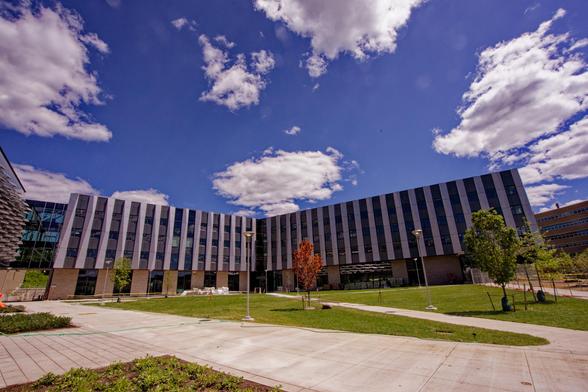Building in a shape a bit like an "L" but with a more than 90 degree angle which notably has alternating light and dark stripes of stell and glass sections on the upper floors and stone-colored and glass sections of a different width at the base.  In front find a plaza with large concrete wlakways and sections of short cropped grass with a few trees recently olanted and streetlights and behind a blue sky punctuated with white cumulus clouds.
