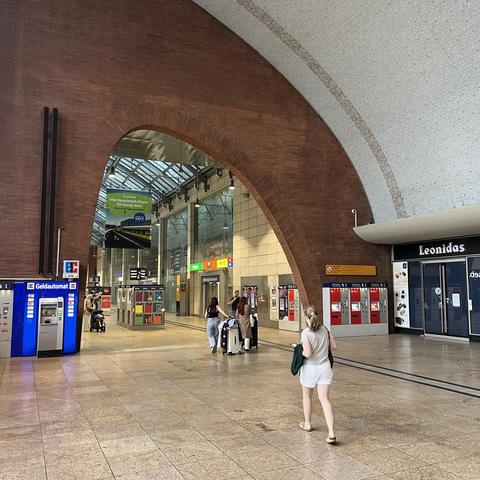 Foto eines Durchgangs im Hauptbahnhof Köln, einige Passant*innen mit Gepäck. Das Dach der Halle und ein geschwungener Backsteindurchgang weisen die typischen Kurven von Möbeln aus den 1950ern auf. Frage mich, wann der Bahnhof gebaut wurde.