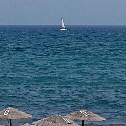 Strahlend blauer Himmel, knapp unterm Horizont zieht ein Einmaster Sportboot mit weißem Segel vorbei, das Meer (-Wasser) kräuselt sich leicht, 
Vordergrund drei Strandschirme mit Schilf?- Halmen gedeckt, haben ein kleines Solarpanel zum Handy - Laden 