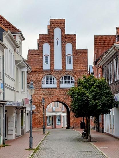 Das Bild zeigt eine historische Brückentorstruktur in einem europäischen Stadtbild. Die Brücke ist aus roten Ziegeln gebaut und hat ein markantes Giebeldach mit drei hohen, weißen Fenstern in der Mitte. Unter dem Tor befindet sich ein Bogen, durch den man in die andere Straßenseite gelangt. Die Straße ist gepflastert und von beiden Seiten von Gebäuden gesäumt, die in verschiedenen Farben und Stilen errichtet sind. Links ist ein weißes Gebäude mit einem Schild "Stoff Paradies" zu sehen, während rechts ein Gebäude mit einem roten Ziegeldach steht. Ein Laternenpfahl mit einer klassischen Laterne steht auf der linken Straßenseite. Vor der Brücke steht ein Baum mit grünen Blättern, der die Szene mit etwas Grün belebt. Der Himmel ist bewölkt, was dem Bild eine ruhige und gedämpfte Atmosphäre verleiht.