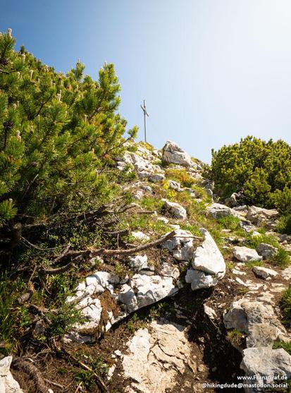 A stunning landscape featuring a rocky hill with a cross on top is depicted in this image. The rocky hill is surrounded by greenery, including trees and bushes, under a clear blue sky. The scene exudes a sense of tranquility and wilderness, with the rocky outcrop and bedrock adding texture to the terrain. This outdoor setting captures the essence of nature, with a mountainous backdrop adding to the picturesque view. The image conveys a serene and peaceful atmosphere, perfect for those seeking s…