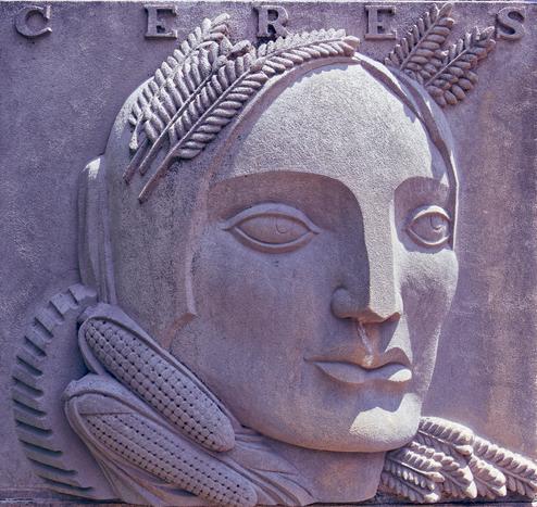 Relief sculpture on stone of a woman's face centered in the frame and looking to the right with long fern-like leaves for hair and ears of corn around her neck and the lettering C E R E S up at the very top