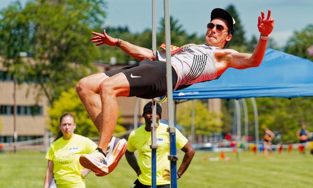 We're looking almost edge-on to the high jump and a man with dark glasses, billed cap and nike shorts seems suspended in the air just about the bar with his arms spread out wide and torso almost horizontal while his legs are bent at the knee at an almost 90 degree angle -- behind him are two volunteers watching who are running the event and a blue tent, a high school building, lots of grass,  pennant flags and two athletes milling around