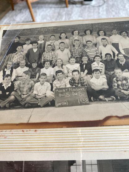 The image is a black and white photograph of a school class from Mount Pleasant, Grade 5, Division 5, taken in 1958. The photograph shows a group of children and adults, likely teachers, arranged in three rows. The front row consists of children sitting on the ground, while the middle and back rows feature children and adults standing. A chalkboard sign in the front center reads "MOUNT PLEASANT. Grade 5. Div 5. (LAYTON) 1958." The children are dressed in typical mid-20th-century attire, with bo…