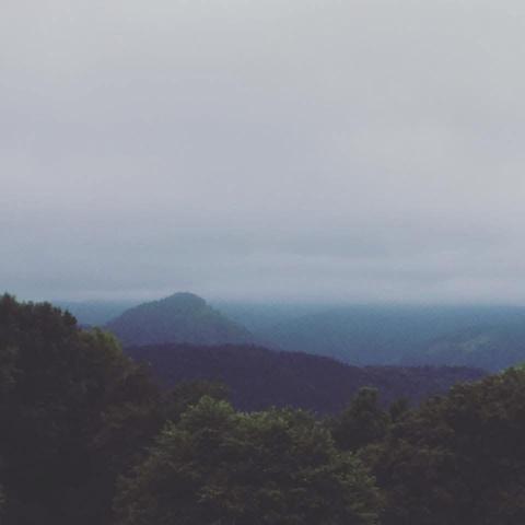 Una cima como una isla entre árboles al frente, colinas y montañas al fondo cubierto de nubes. Un cielo de algodón con un ambiente de ligera neblina. 
