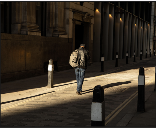 Man wearing leather jacket and blue jeans, with a rucksack slung over his left shoulder, walking away from the camera. He is walking between two rows of bollards in a low beam of sunlight; the rest of the street is in shadow.