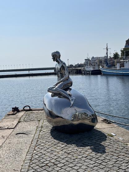 Silbrig verspiegelte Statue eines sitzenden Knaben (a la kleine Meerjungfrau) im Hafen von Helsingør, der Richtung Schweden guckt. Meer und Kaianlage.