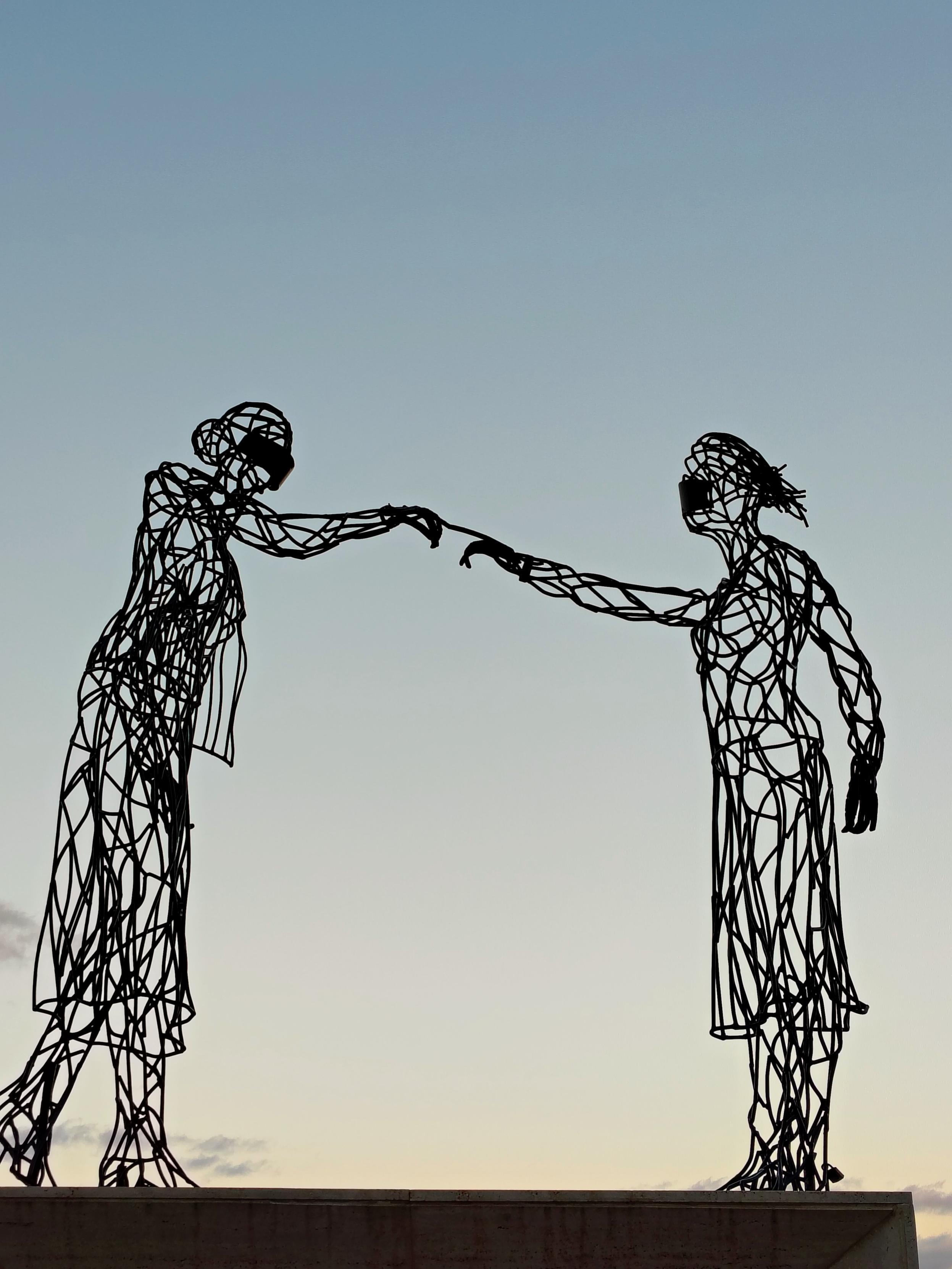 Man sieht ein Denkmal bestehend aus zwei weiblichen Metallskulpturen. Sie tragen Mundschutz und berühren sich nur mit jeweils einem Finger. Man sieht ein Denkmal bestehend aus zwei weiblichen Metallskulpturen. Sie tragen Mundschutz und berühren sich nur mit jeweils einem Finger.