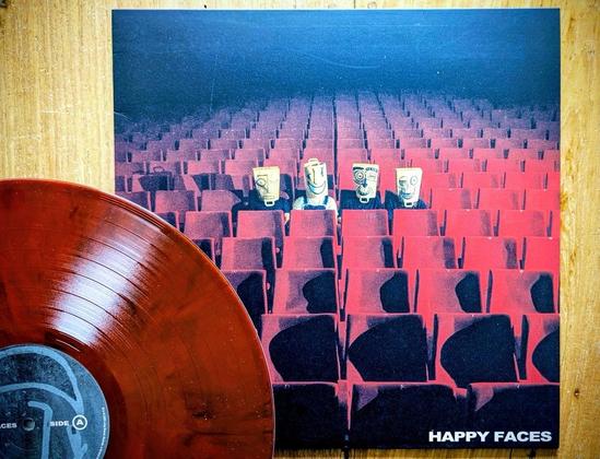 Das Bild zeigt eine Schallplatte mit rötlich-braunem Cover. Das Artwork zeigt eine Reihe leerer roter Theatersitze, in deren Mitte vier Zeichentrickfiguren mit Masken und fröhlichen Gesichtern Platz nehmen. Der Titel "HAPPY FACES" ist auf dem Cover zu lesen.