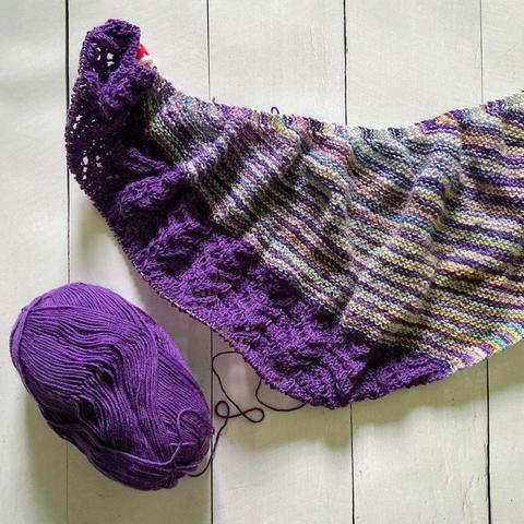 Flat-lay view of an asymmetrical shawl project on a circular needle in garter stitch, it has stripes in various pastel colors, and a couple of repeats of a lace border in solid purple. The ball of yarn lies is also there on the white plank background.