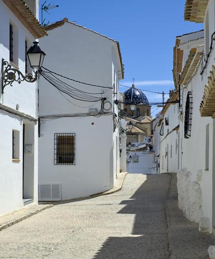 Fotografía de una calle solitaria al mediodia entre casas de paredes blancas, en el lado derecho de la calle hay sombra en el izquierdo luz, al fondo de la calle se ve la cúpula azul de la iglesia del pueblo.