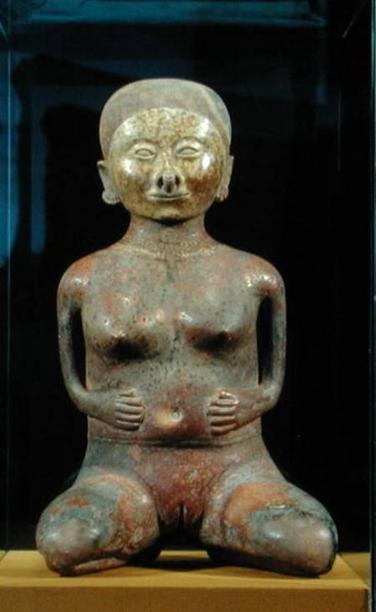 Statuette of a woman kneeling with her legs apart enough for the onlooker to see a vulva with slit. Her hands are placed on her belly and she as small breasts.