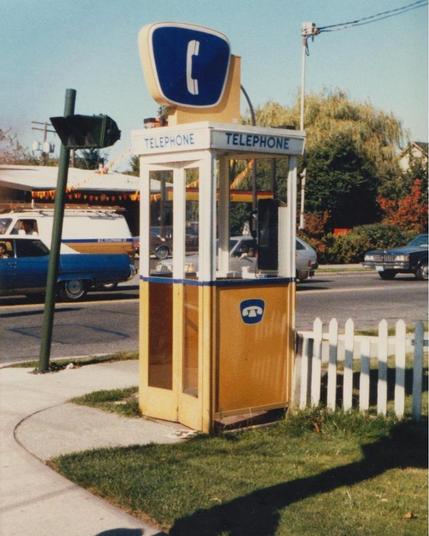 The image depicts a vintage public telephone booth situated on a sidewalk. The booth is painted in a bright yellow color with white trim and features a large blue sign with a white telephone icon and the word "TELEPHONE" written in blue letters. The booth has a glass front and two doors, with a small window visible on the side. A white picket fence surrounds the booth, and a grassy area is visible in front of it. In the background, there is a street with vehicles, including a blue and white sta…