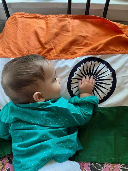 A baby wearing a turquoise kurta with yellow dots is lying on an Indian flag, featuring orange, white, and green colors. The baby is reaching towards the chakra in the center of the flag.