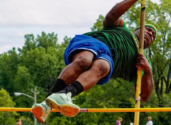 Large black man looks like he's succeeding at the pole vault as he holds on to his pole as his feet go over the bar;  he's wearing a green short,  blue shorts and a green and yellow headband.