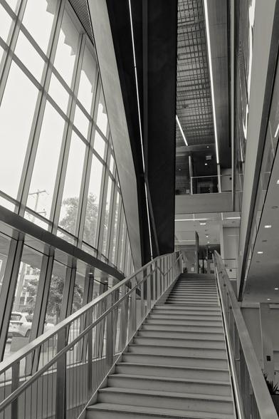 Monochrome Image of two stairways going in opposite directions -- on the right side a staircase rises up halfway away from me,  then another staircase heads up on the left back to my side;  on the left of it all you can see a street scene through  multiple-paned windows.