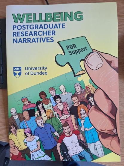 Picture of a comic cover with a a group of people looking at or in the rough direction of the viewer. They are diverse in many ways, e.g. clad differently, of different ages (although most looking young), different skin tones, at the front is someone with a wheelchair. Their picture seems to be a puzzle, with a big hand coming in and putting the last piece in the top right corner (showing the words "PGR Support"). The comic is titled "Wellbeing - Postgraduate Researcher Narratives", and branded "University of Dundee"