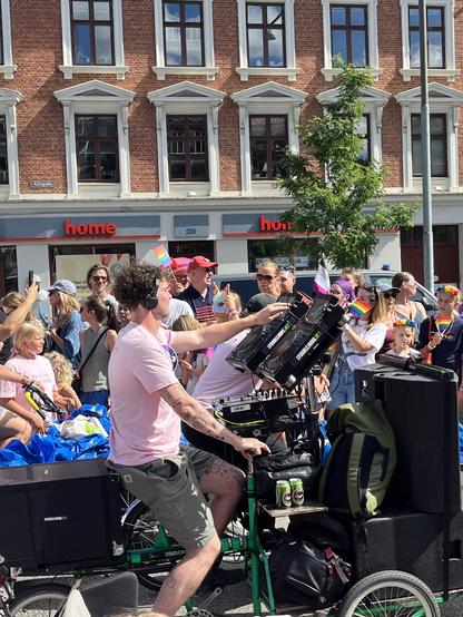 Ein DJ bedient ein Mischpult und fährt gleichzeitig ein Lastenrad mit Box. Im Hintergrund ein Gebäude und Menschenmenge am Straßenrand.