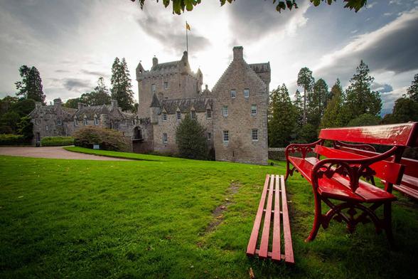 A serene outdoor scene featuring a red bench positioned in front of a majestic castle, with Cawdor Castle visible in the background. The lush green grass and clear blue sky create a peaceful atmosphere, with a few trees and a palm tree adding to the natural beauty of the landscape. The castle, with its grey stone walls and turrets, stands tall and imposing against the sky. The wooden bench, painted in a bright red color, offers a spot for relaxation and contemplation in this picturesque park se…