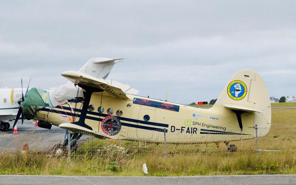 A faded yellow propeller bi-wing plane is parked in the grass. It has a cover over the windshield and various markings, including the registration D-FAIR