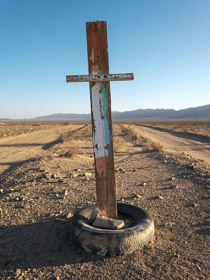 Crude wooden cross with tire base standing at a slant between two parallel dirt roads in desert landscape.