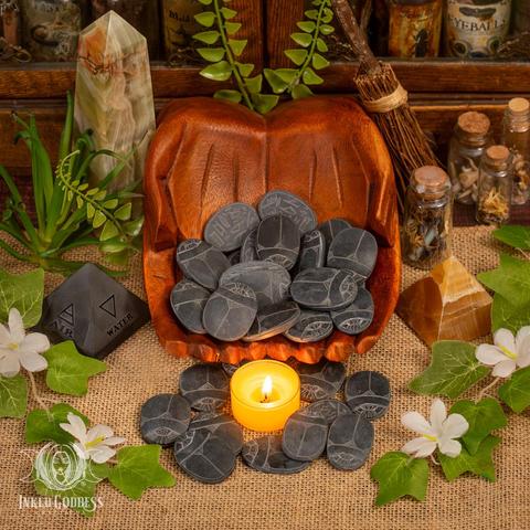 Several Basalt Carved Scarabs rest in cupped, carved wooden hands, while other specimens are arranged on a burlap-covered surface. They are dark grey, relatively flat and oval, and a little smaller than a tealight candle, shown for scale. The carving on them stands out as a lighter grey; on top is a stylized scarab beetle, and the bottom features hand-carved Egyptian hieroglyphics. Presented by Inked Goddess Creations.