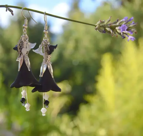 A pair of black flower earrings, dangling by their hooks from a stem of lavender. White and black crystals dangle on stamens beneath, and smaller flowers flank the central larger flower. Blurred greenery and sunshine behind.