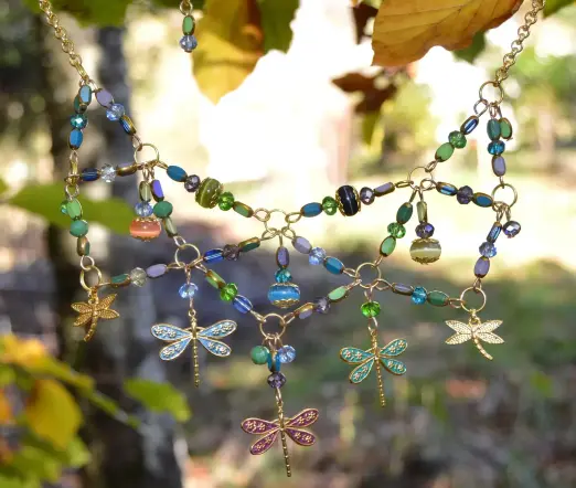 Beaded necklace with colourful dragonflies, against a blurry autumn tree. The bib shape is made of zigzag beaded sections, fanning out, bead pendants hanging from the links. The glass beads are various blues, light purples and greens, with gold edges.