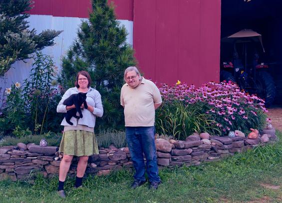 Towards the center left find a woman wearing a green knee-length skirt and a blue shirt holding a large black vat and next to her a man with grey hair and a beige shirt and blue jeans.  Behind them a stone wall,  two evergreen trees,  a few lily flowers to the left,  and many purple coneflower to the right,  and behind that a red barn wall with a blue stripe painted on it, through the open door you might make out a blue tractor
