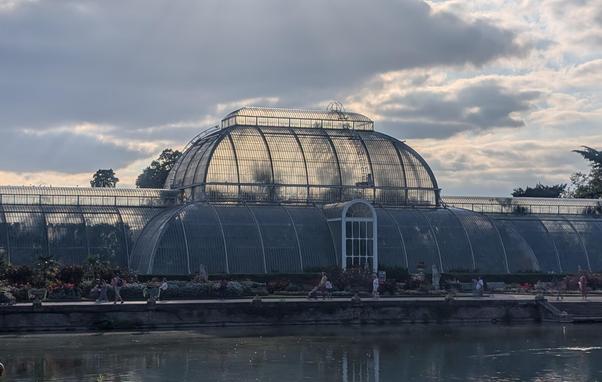 A wide shot of a grand, multi-sectioned glass and metal conservatory, likely a greenhouse, under a cloudy sky. The building dominates the upper half of the image, featuring a large, prominent central dome and long, arched sections extending to either side. Sunlight reflects brightly off the glass panes of the central dome. In the foreground, a wide pond or lake stretches across the bottom of the image, reflecting the sky and parts of the building. Between the water and the conservatory, a paved walkway is visible, lined with lush flowerbeds and potted plants, where several people are seen strolling or standing. Trees are visible in the background on either side of the conservatory.