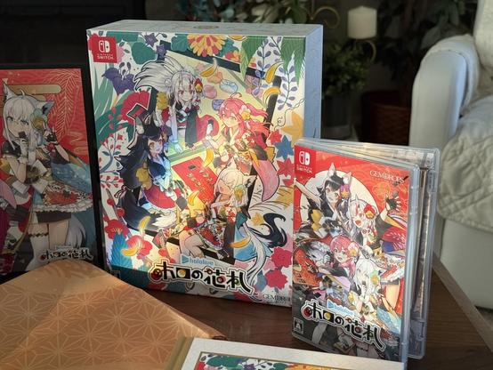 A closeup of the box for Holo’s Hanafuda Exclusive Hololive Edition and the game box for Nintendo Switch. The art on the cardboard box features four of the members sitting around a mat playing hanafuda. The game box shows the same four members with hanafuda-related icons around them. 