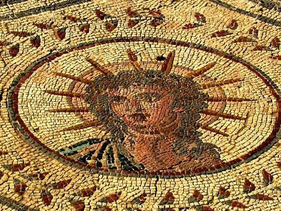 Mosaic of Helios as part of the Mosaic of the Planetarium. We see his bust inside a circle, decorated by an outer circle of decorative leaves. Helios wears a dark fabric on his right shoulder, likely a chlamys cloak. His curly hair falls to his shoulders and his head is illuminated by an aureola with 12 spikes.