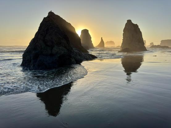 Silhouetted rock formations rise from a sandy beach at sunset, reflecting in the wet sand. Gentle waves lap at the shore, and a soft golden light creates a serene atmosphere.