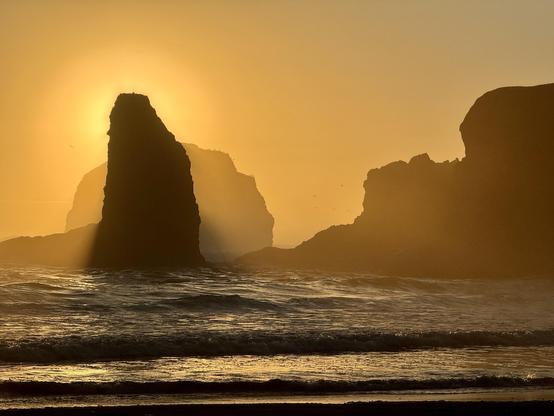 A dramatic sunset casts a golden light over a beach scene, highlighting silhouetted rock formations rising from the ocean waves. The soft glow creates a misty atmosphere, with birds faintly visible in the sky.