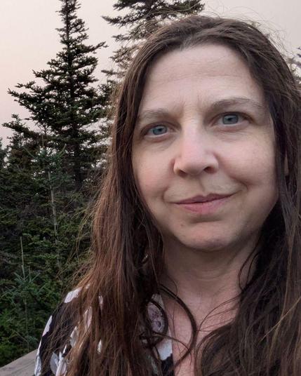 Selfie of a white woman with blue eyes and long brown hair wearing a floral top. She's standing on a brown deck and sort of smirking. Perhaps she is bemused. Behind her are some evergreen trees, and a smokey pinkish coloured sky.